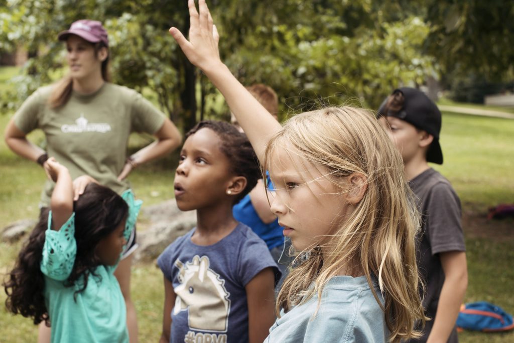 Young blonde girl raising hand surrounded by other children and camp counselor